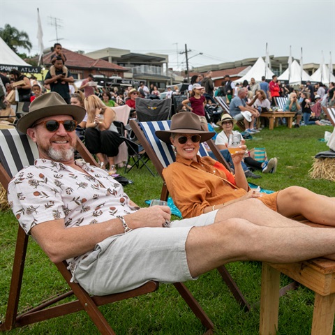 Two people lying on deck chairs with glasses of wine with crowd behind