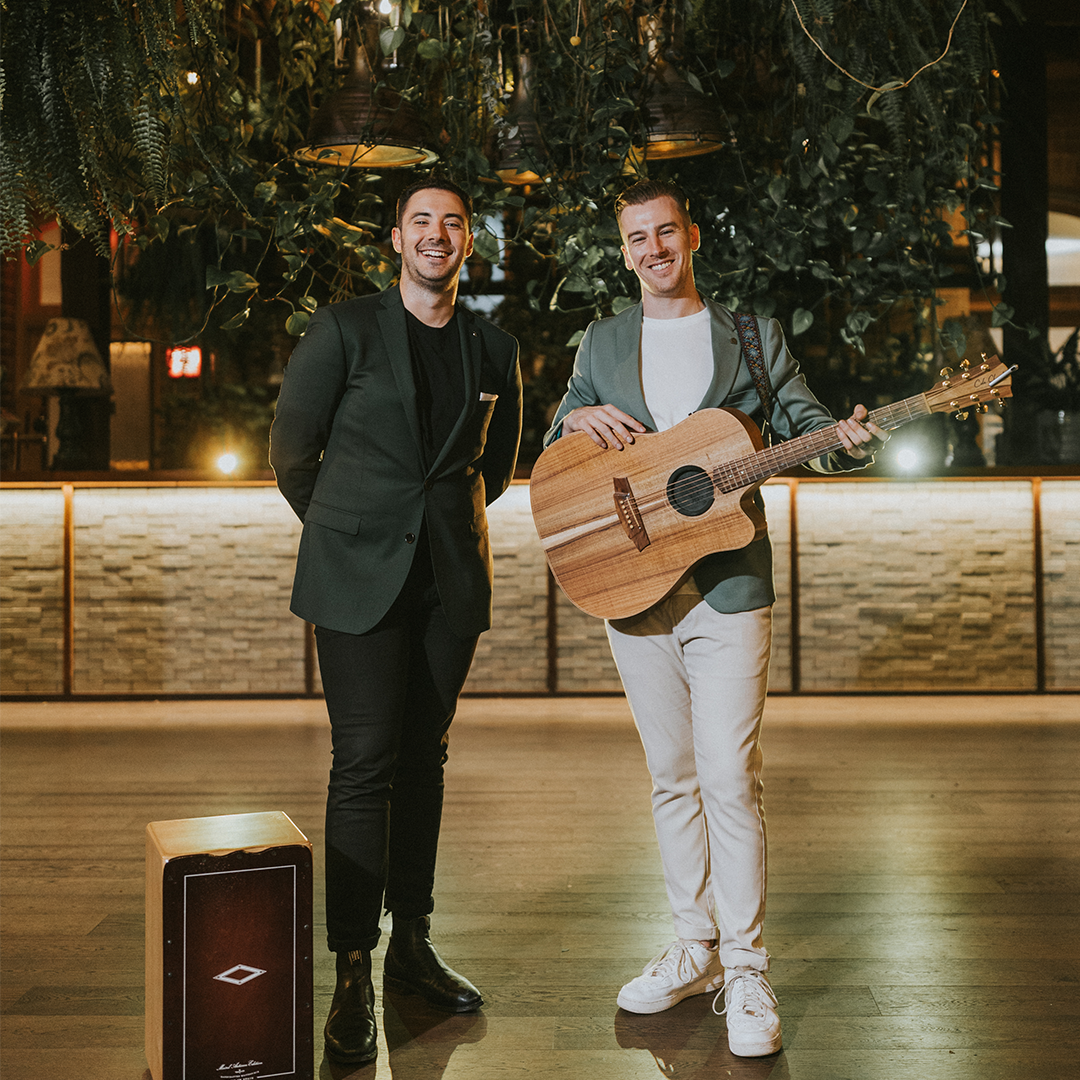 Two men pose for the camera, they have an acoustic guitar and cajon