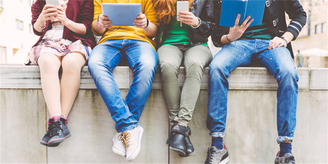 Four young sitting on stone wall, seen from the shoulders down, they each hold a device or book.