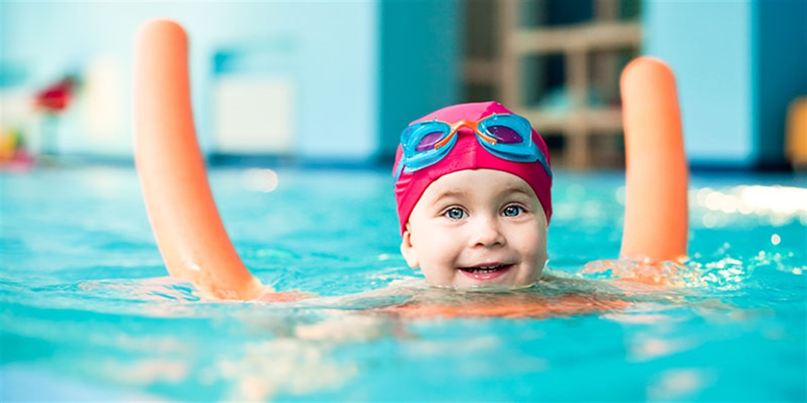 Baby wearing pink cap and goggles swimming in pool using floating device