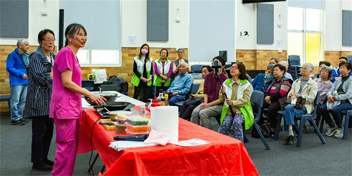 Two ladies stand at a table with food and hotplate, giving a cooking class to a seated audience