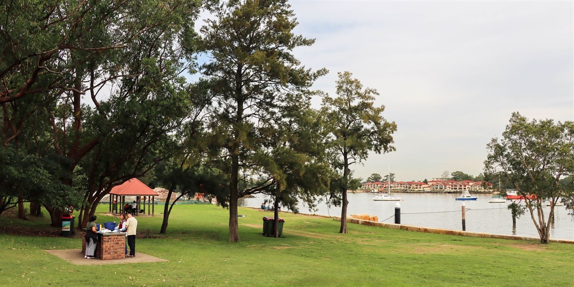 Putney Park with people using the BBQs on a grass lawn with leafy tress behind them and the Parramatta River in the background.