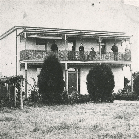 Old photo of a double storey house, with 5 adults standing on the balcony