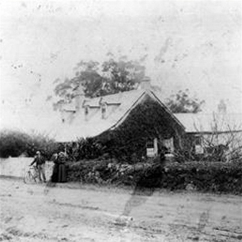 An old couple stands by the entrance of a house. A plaque monuments sits on the nature strip.
