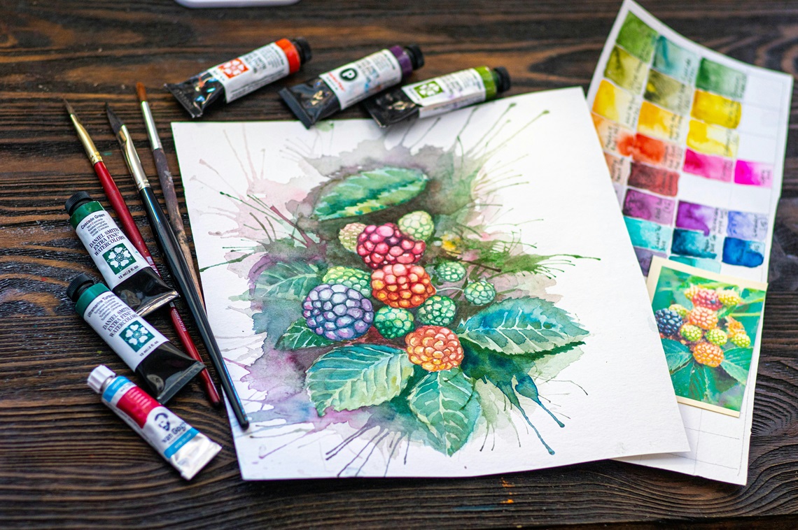 View of a watercolour painting of leaves and berries, sitting on a wooden table, surrounded by brushes and paint tubes