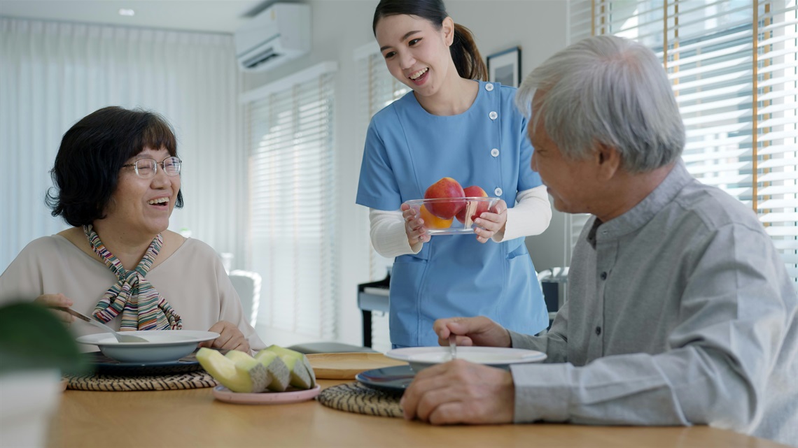 Two seniors, man and woman, sit at a table. An aged care worker brings them a bowl of fruit.