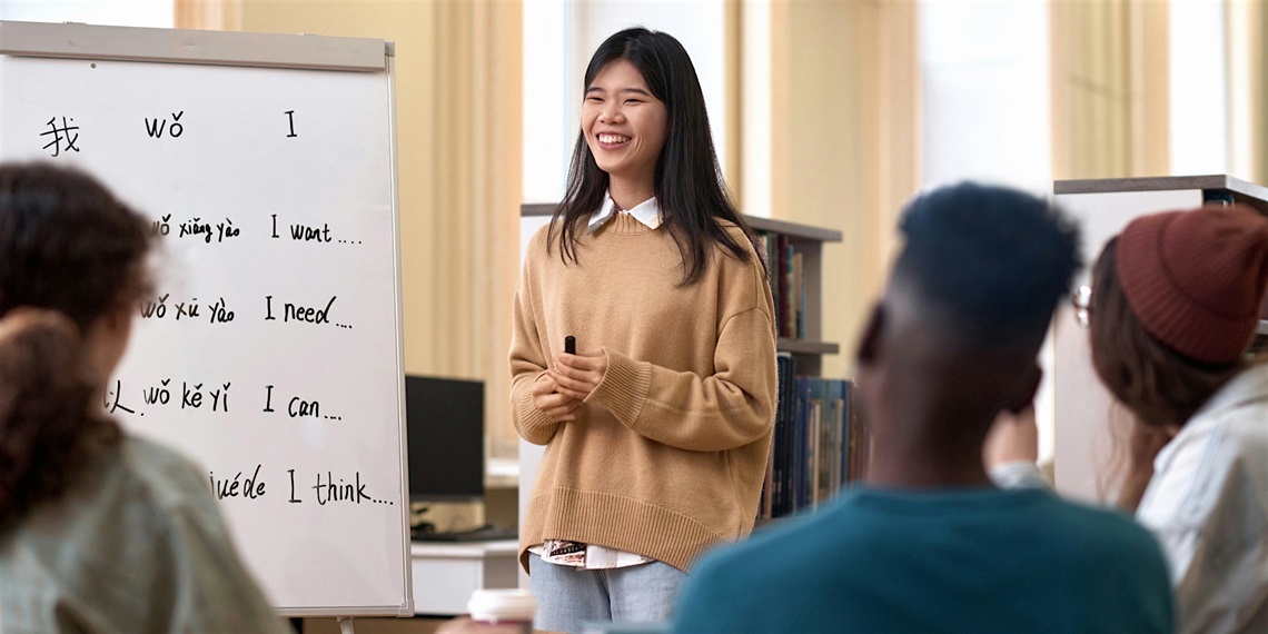 Female teacher stands by a whiteboard, in front of three seated students