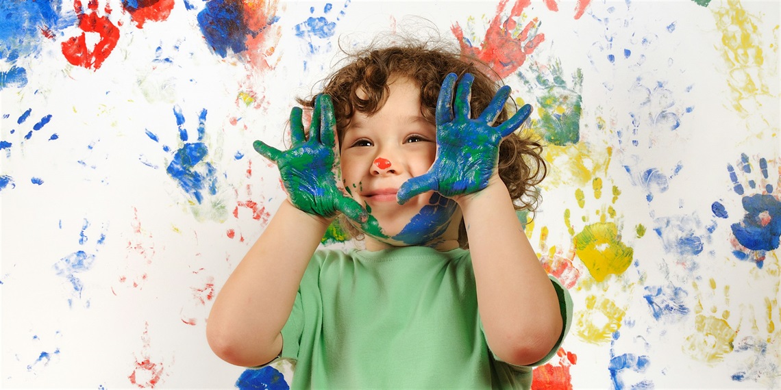 Child with curly hair holds up hands covered in paint. There are paint handprints on the wall behind her.
